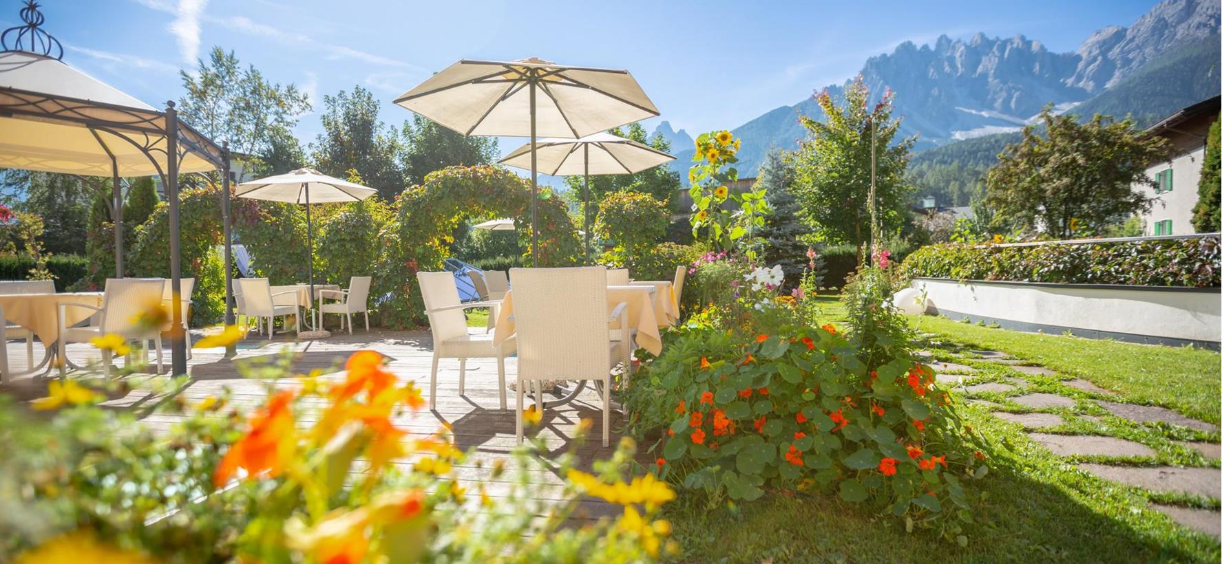 Terrasse der Villa Stefania mit weißen Stühlen und Tischen umgeben von bunten Blumen und den Dolomiten im Hintergrund.