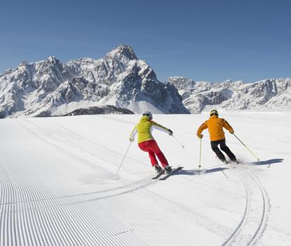 Two skiers on a groomed ski slope in the snow-covered mountain landscape of the Dolomites.