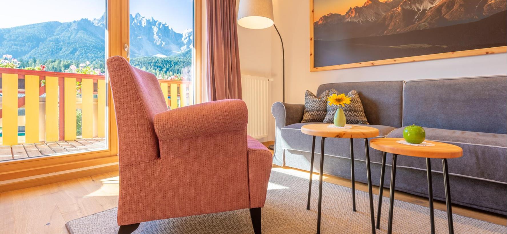 Sitzecke der Suite Dolomiti mit grauem Sofa, orangen Sesseln und Blick auf den Balkon und die Dolomiten.