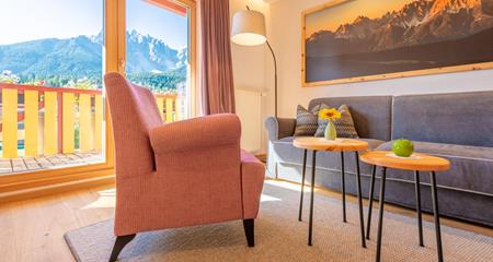 Sitzecke der Suite Dolomiti mit grauem Sofa, orangen Sesseln und Blick auf den Balkon und die Dolomiten.