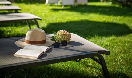 Lettino prendisole con cappello e libro nel verde giardino di Villa Stefania, con alberi e le cime delle Dolomiti sullo sfondo.