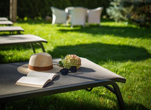 Lettino prendisole con cappello e libro nel verde giardino di Villa Stefania, con alberi e le cime delle Dolomiti sullo sfondo.
