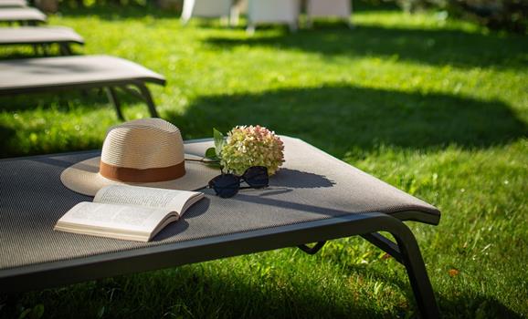 Sun lounger with a hat and book in the green garden of Villa Stefania, with trees and the Dolomite peaks in the background.