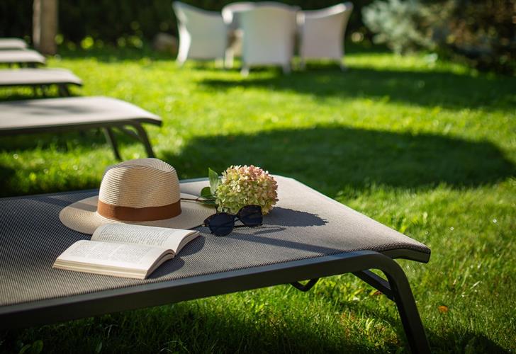 Sonnenliege mit Hut und Buch im grünen Garten der Villa Stefania mit Bäumen und Dolomitengipfeln im Hintergrund.