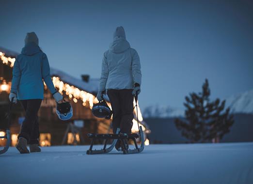 Zwei Personen in Winterkleidung ziehen einen Schlitten; daneben ein beleuchtetes Gebäude nachts..