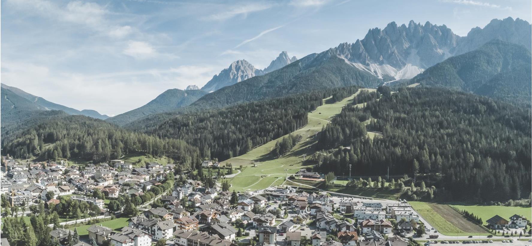 Luftaufnahme von Innichen mit Wiesen, Wäldern und Bergen im Hintergrund.