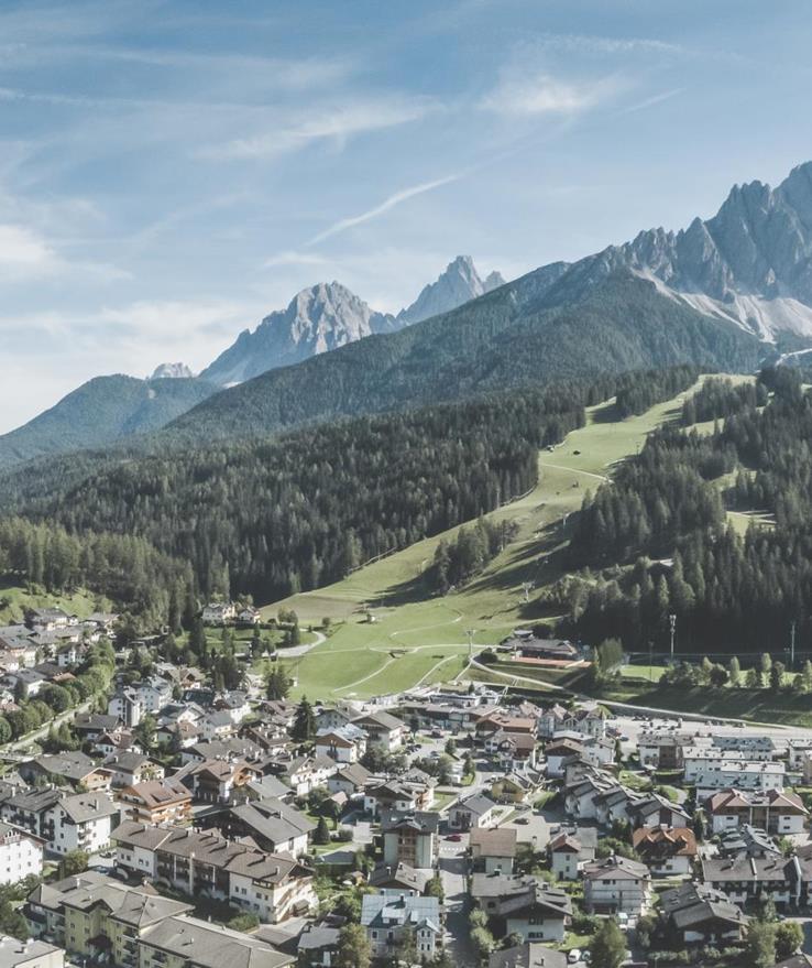 Luftaufnahme von Innichen mit Wiesen, Wäldern und Bergen im Hintergrund.