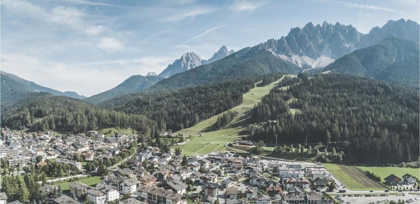 Vista aerea di San Candido con prati, boschi e montagne sullo sfondo.