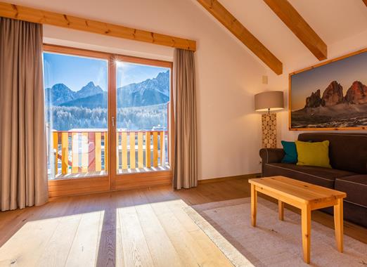 Living area of the Natura Suite with sofa, exposed wooden beams and balcony with panoramic views of the Dolomites.