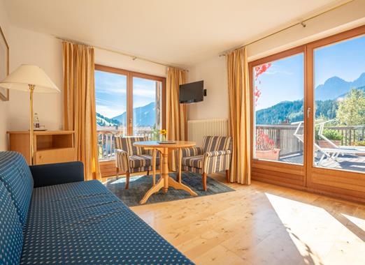 Soggiorno della Junior Suite con divano blu, angolo salotto accanto alla finestra e balcone con vista sulle Dolomiti.