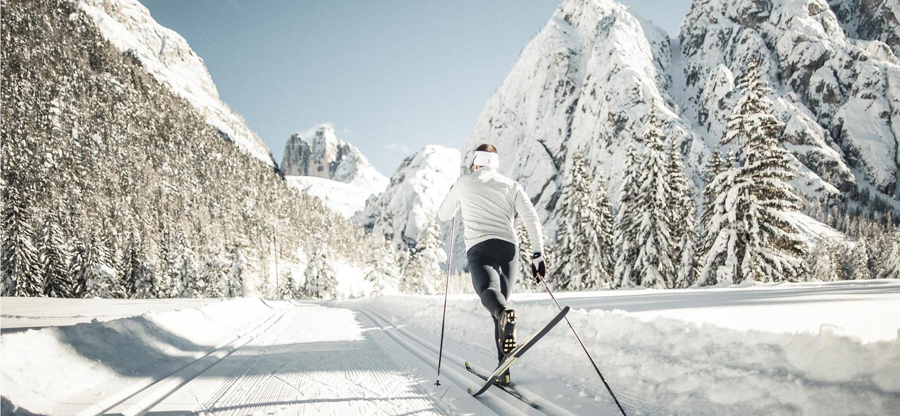 Ein Langläufer auf einer Loipe in Winterlandschaft mit schneebedeckten Bäumen und Bergen im Hintergrund.