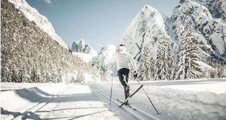 Ein Langläufer auf einer Loipe in Winterlandschaft mit schneebedeckten Bäumen und Bergen im Hintergrund.