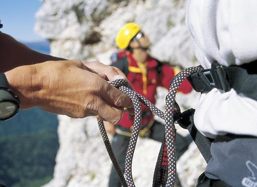 Im Vordergrund Hände die ein Kletterseil halten; im Hintergrund eine weitere Person mit Helm an einer Felswand.