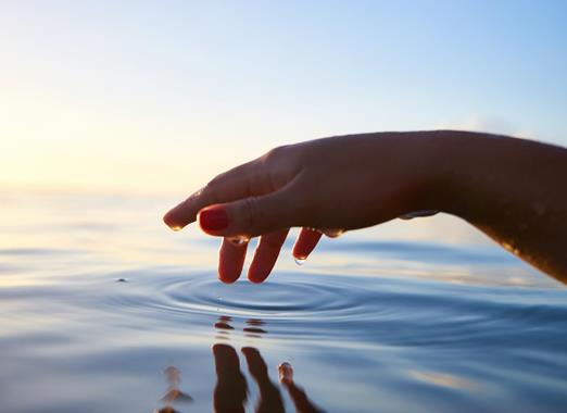 Primo piano di una mano sopra una superficie d’acqua calma al tramonto.