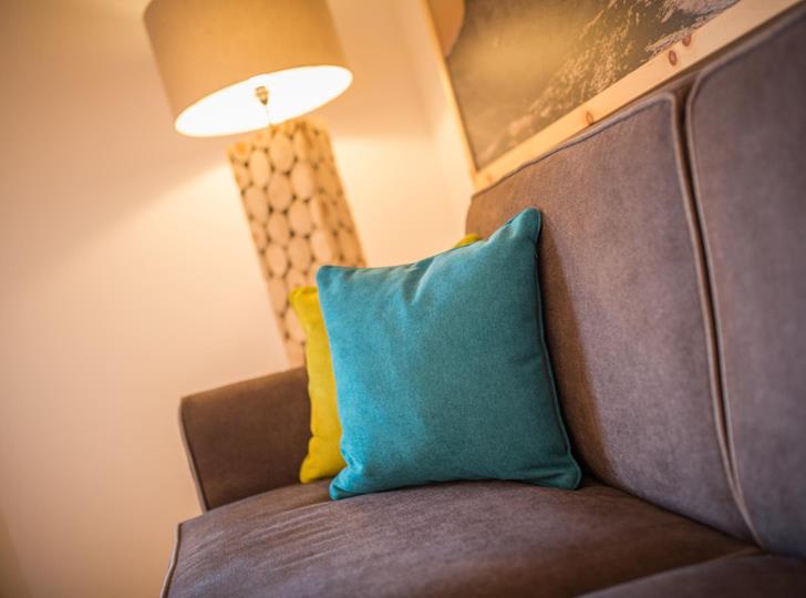Gemütliche Sitzecke mit grauem Sofa, türkisfarbenem Kissen, warmer Beleuchtung und dekorativer Wandgestaltung.