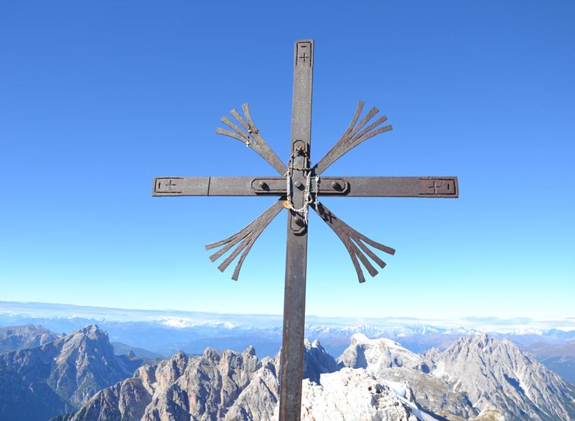 Gipfelkreuz mit Bergpanorama in den Dolomiten und klarem blauem Himmel im Hintergrund.