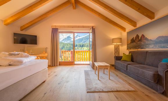 Suite Natura with double bed, sofa, exposed wooden beams and large windows overlooking the mountain landscape of the Dolomites.