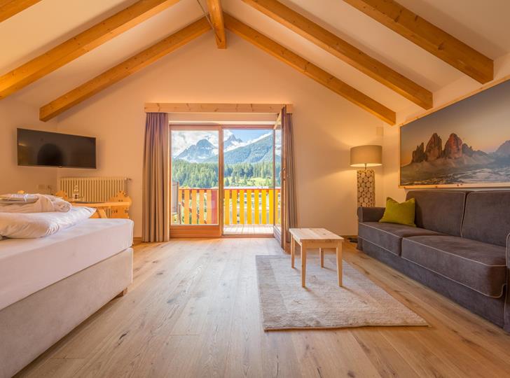 Suite NaturaSuite Natura con letto matrimoniale, divano, travi in legno a vista e ampia vetrata con vista sul paesaggio montano delle Dolomiti.