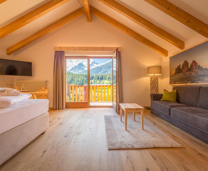 Suite NaturaSuite Natura con letto matrimoniale, divano, travi in legno a vista e ampia vetrata con vista sul paesaggio montano delle Dolomiti.