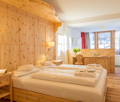Romantic Suite rivestita in legno con letto matrimoniale, asciugamani sul letto, angolo salotto e finestra con vista sulle Dolomiti.
