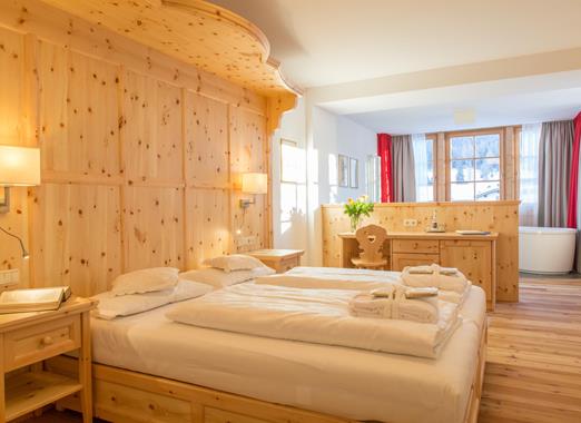 Romantic Suite in Holz mit Doppelbett, aufgelegten Handtüchern, Sitzecke und Fenster mit Blick in die Dolomiten.