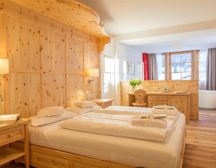 Romantic Suite in Holz mit Doppelbett, aufgelegten Handtüchern, Sitzecke und Fenster mit Blick in die Dolomiten.