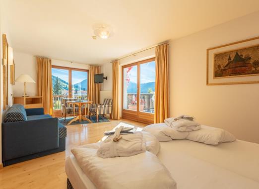 Junior Suite con letto matrimoniale, angolo salotto con divano blu e poltrona e porta-finestra del balcone con vista sulle montagne.